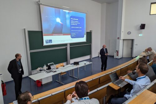 Ein Vortragender präsentiert vor Publikum in einem Hörsaal, während eine Präsentationsfolie auf einer großen Leinwand angezeigt wird.