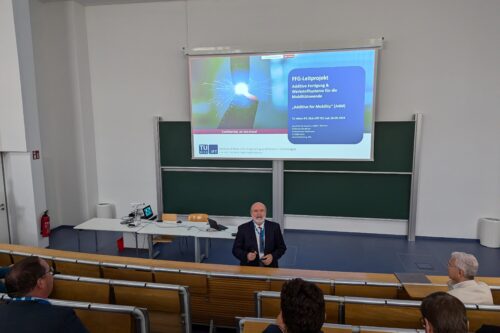 Ein Vortragender spricht vor einem sitzenden Publikum in einem Hörsaal, begleitet von einer Präsentation auf der Leinwand.