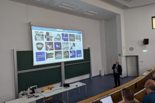 Ein Vortragender zeigt eine Präsentationsfolie mit verschiedenen Themenfeldern der Produktionstechnologie in einem Hörsaal.