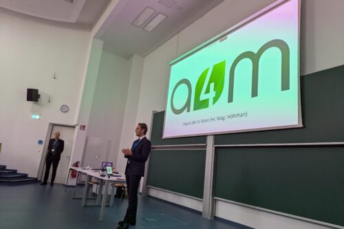Ein Referent hält einen Vortrag vor einer großen Leinwand mit der Aufschrift „a4m“ in einem Hörsaal.