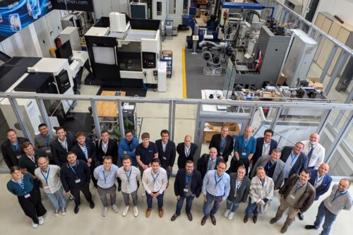 Eine größere Gruppe von Teilnehmenden steht für ein gemeinsames Foto in einer Produktionshalle mit Maschinen im Hintergrund.