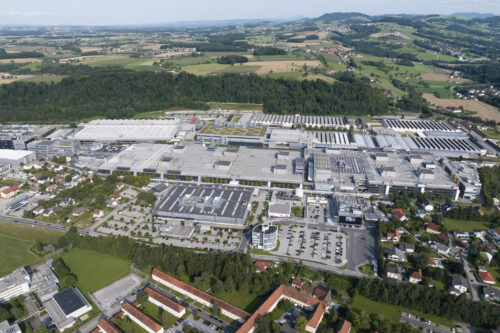 Aerial view of a large industrial and production site surrounded by residential areas, fields, and a hilly landscape.
