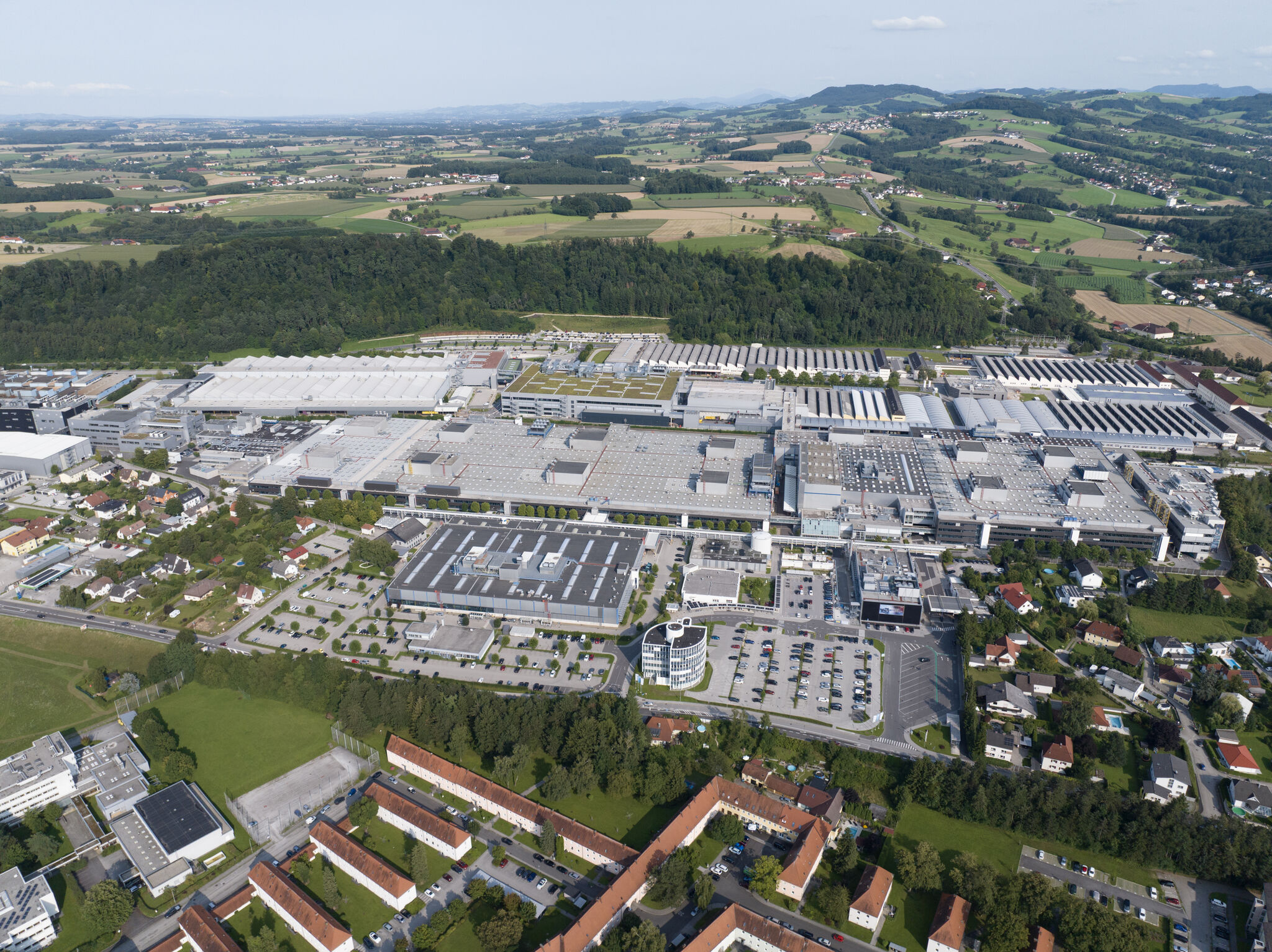 Aerial view of a large industrial and production site surrounded by residential areas, fields, and a hilly landscape.
