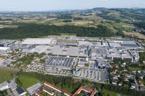Aerial view of a large industrial and production site surrounded by residential areas, fields, and a hilly landscape.