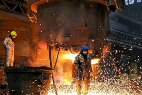 Two foundry workers in heat-protective clothing work amid flying sparks on a glowing, large-scale steel casting.
