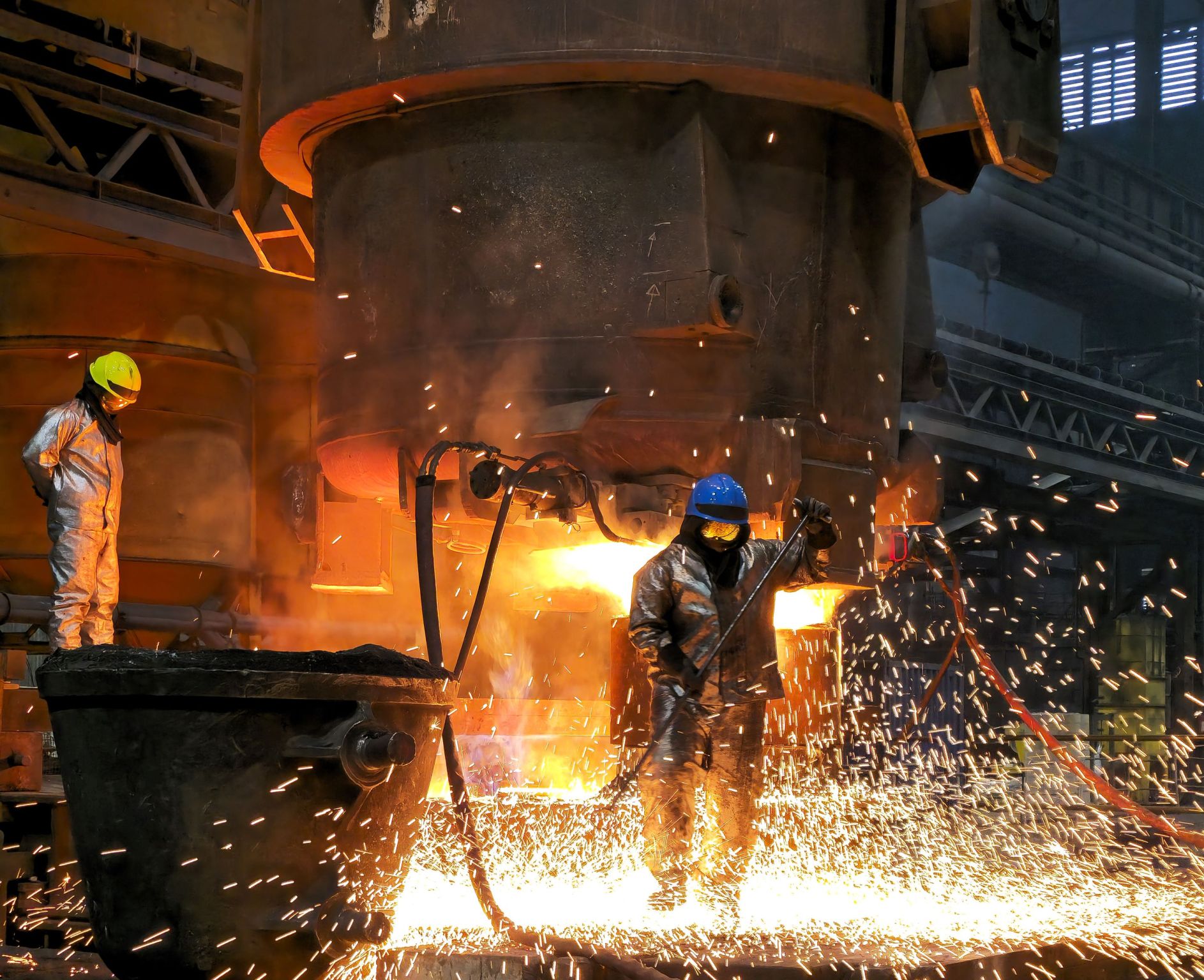 Two foundry workers in heat-protective clothing work amid flying sparks on a glowing, large-scale steel casting.