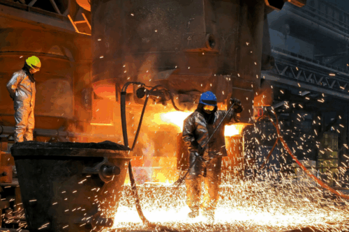 Two foundry workers in heat-protective clothing work amid flying sparks on a glowing, large-scale steel casting.